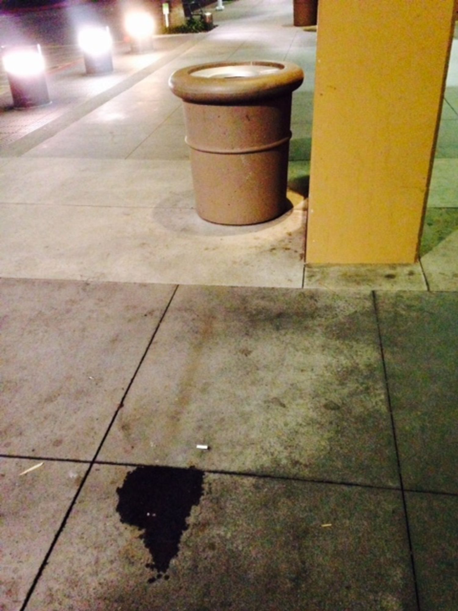A brown trash can sits on a sidewalk next to a pillar