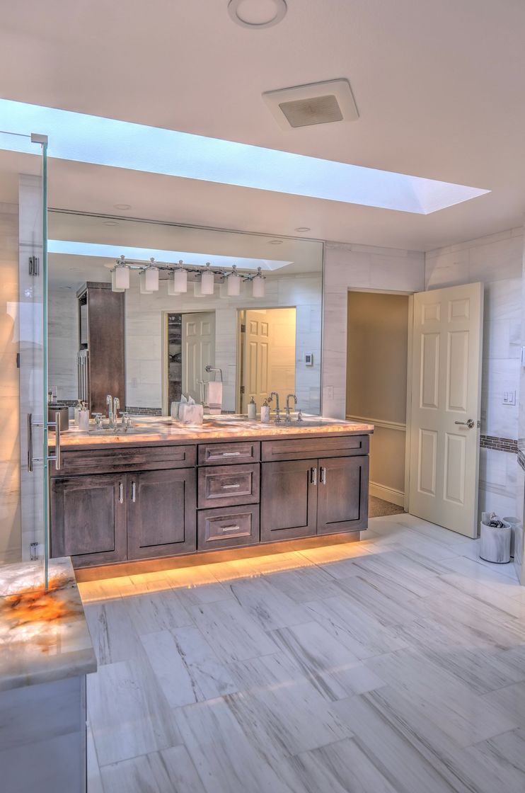 Bathroom with a double sink vanity, large mirror, and skylight. Light wood cabinets and marble flooring.