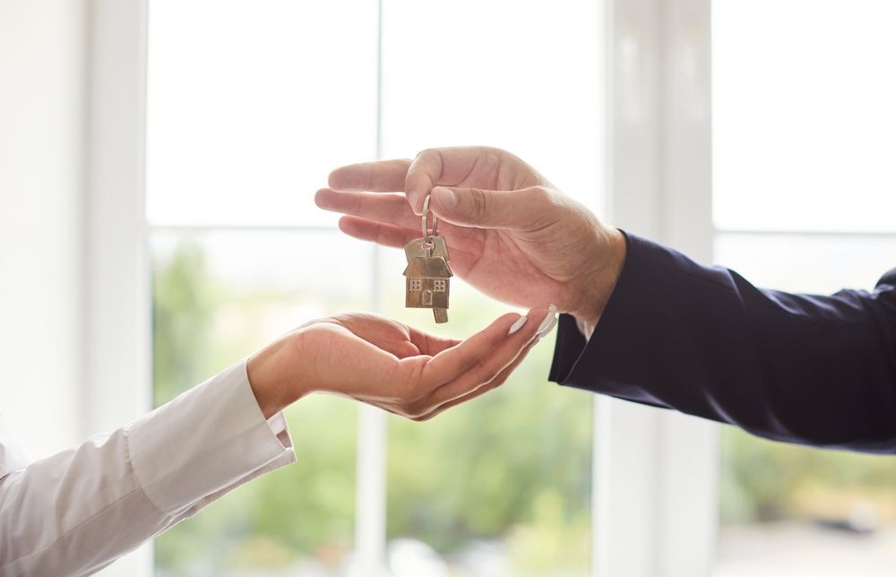 One person in a suit hands a small house-shaped key to another person against a bright, out-of-focus window background.