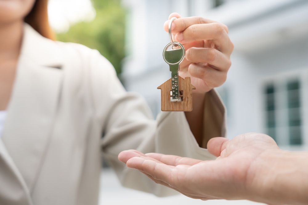 A person in a light-colored blazer hands a house-shaped keychain with a metal key to another person.