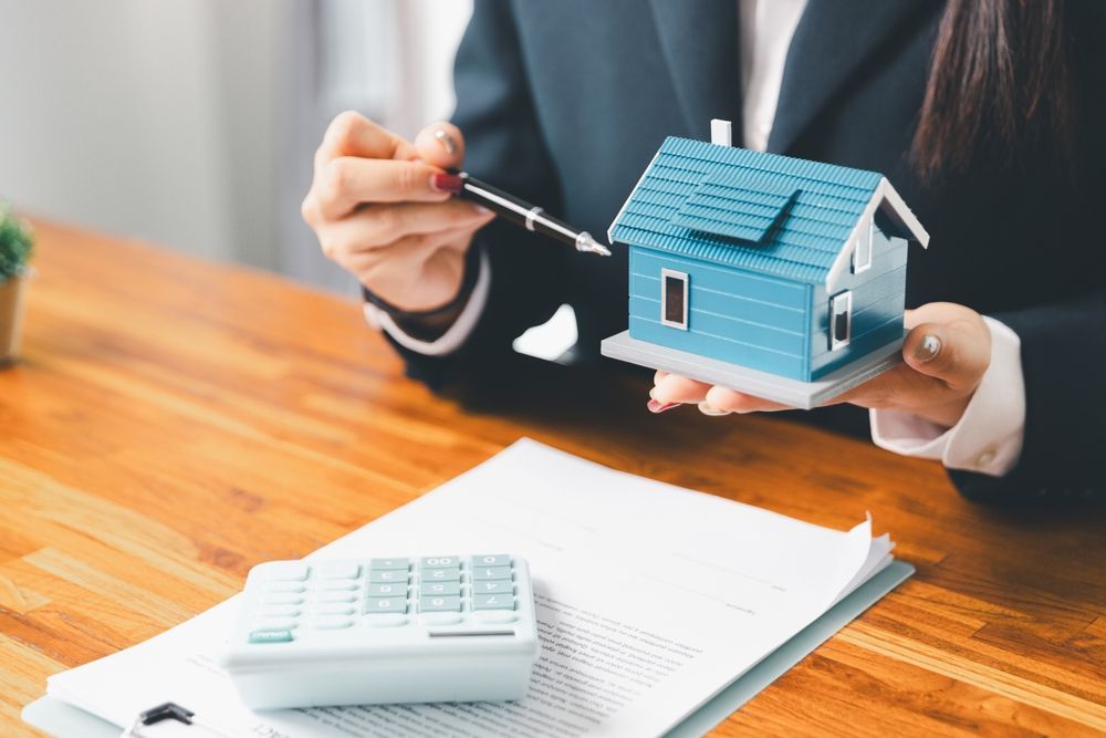 A person in a business suit holding a small blue model house and pointing with a pen, with a calculator on a document.