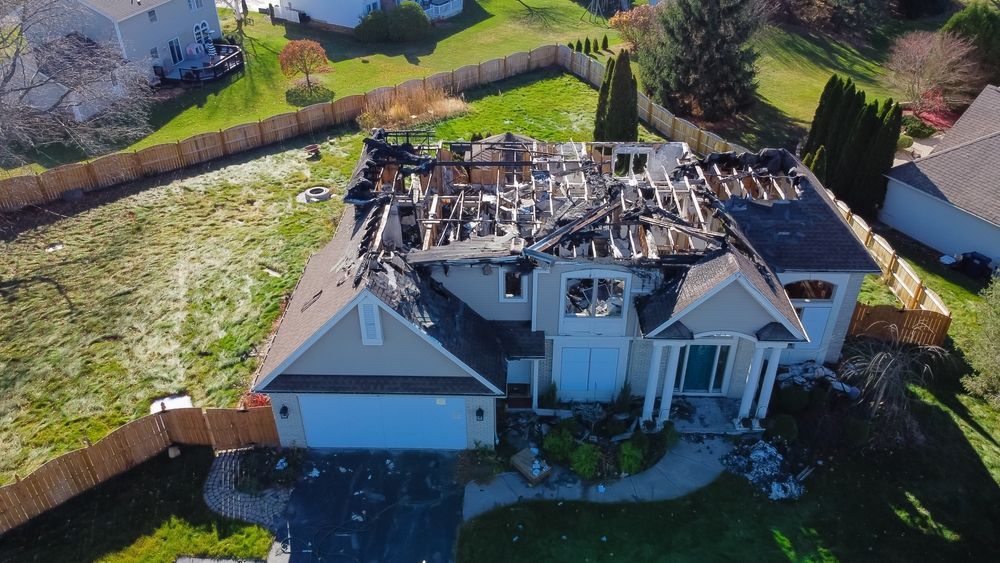 Aerial view of a residential house with a partially destroyed, charred roof due to fire damage in a suburban yard.