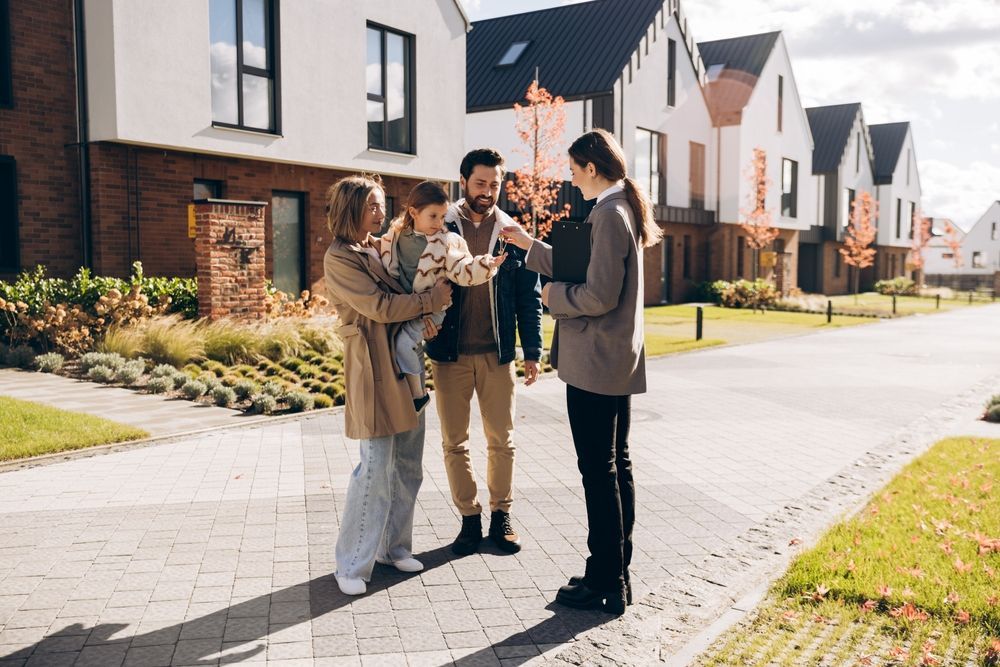 A real estate agent hands keys to a family outside a row of modern, newly constructed suburban houses.