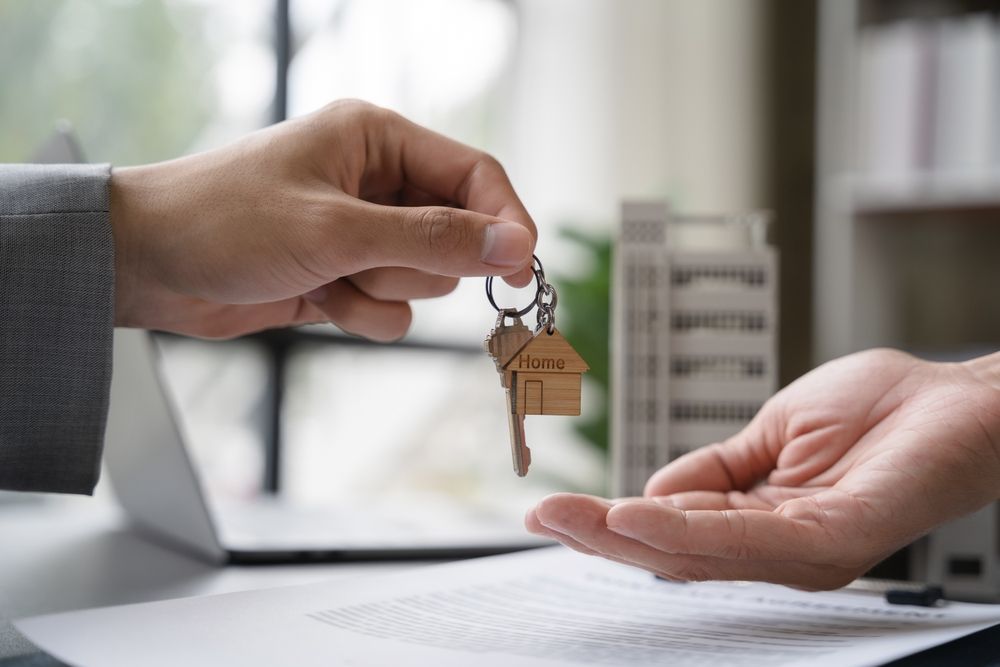 A person in a business suit hands keys with a house-shaped keychain to another person over a contract.