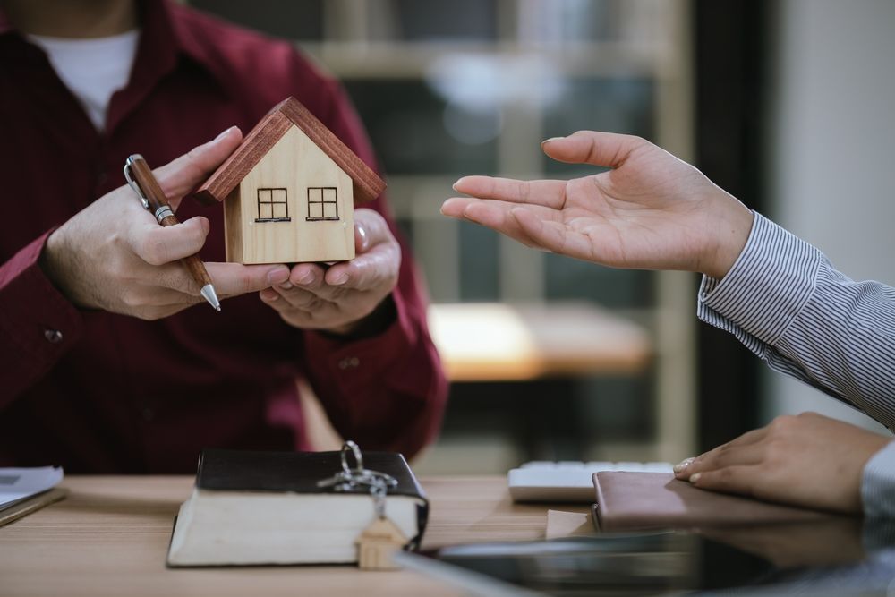 Two people at a desk, one offering a small wooden house model while the other gestures, suggesting a real estate meeting.