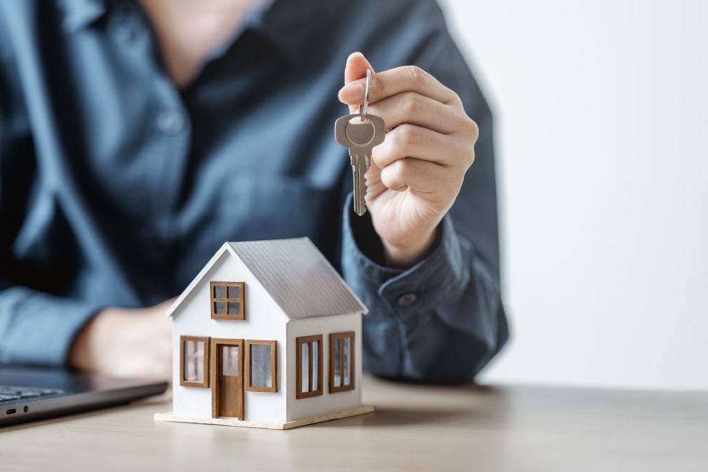 A person in a blue shirt holding a house key above a small model house on a desk.