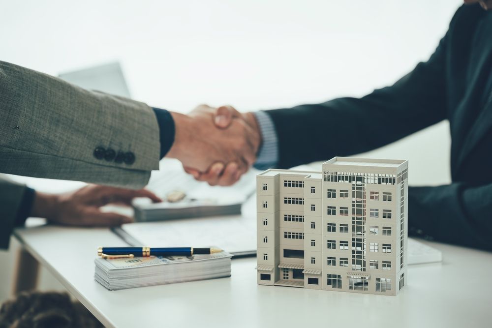 Two professionals shake hands over a table featuring a model of a building and a stack of currency.