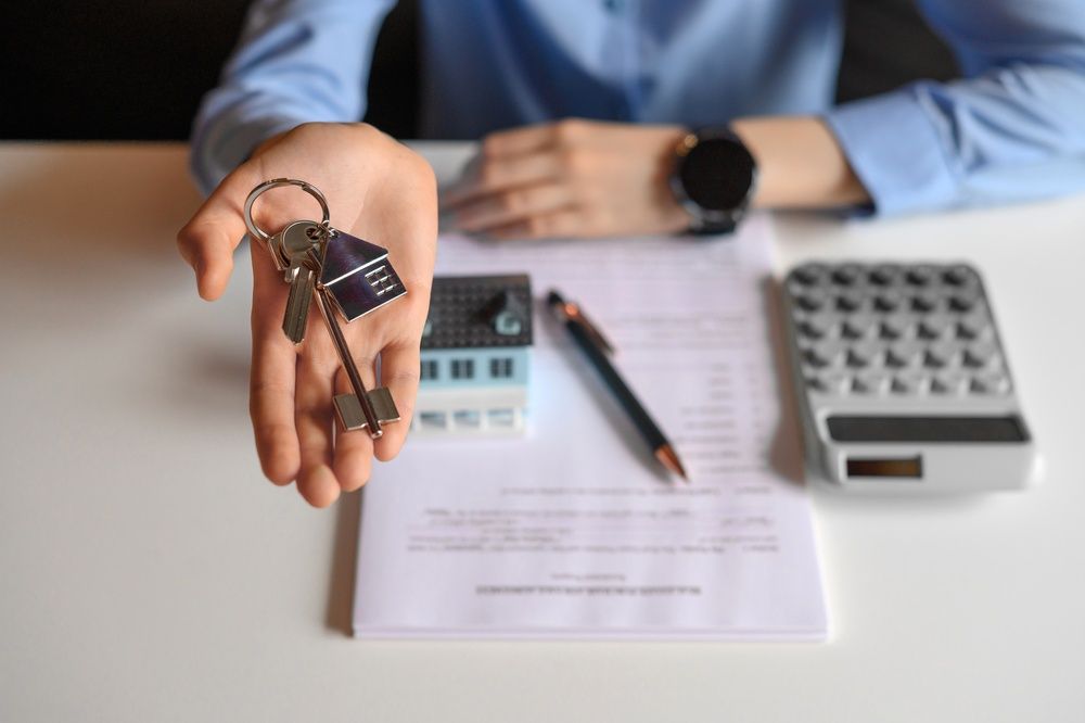 A person in a blue shirt holds house keys with a keychain over a document, a model house, a pen, and a calculator on a desk.