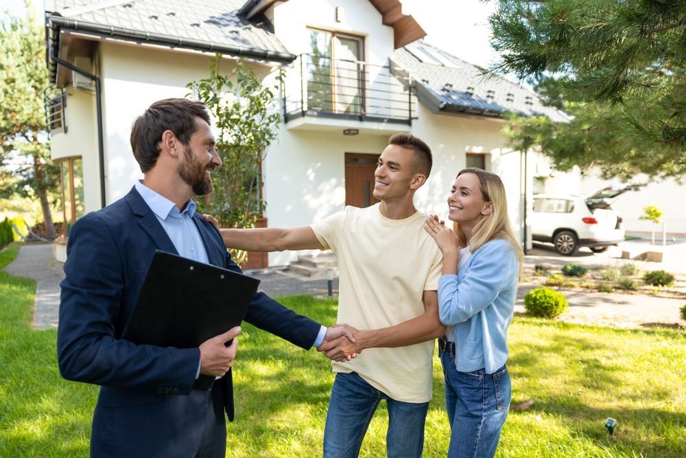 A realtor shakes hands with a couple in front of a house, appearing friendly and professional outdoors on a sunny day.