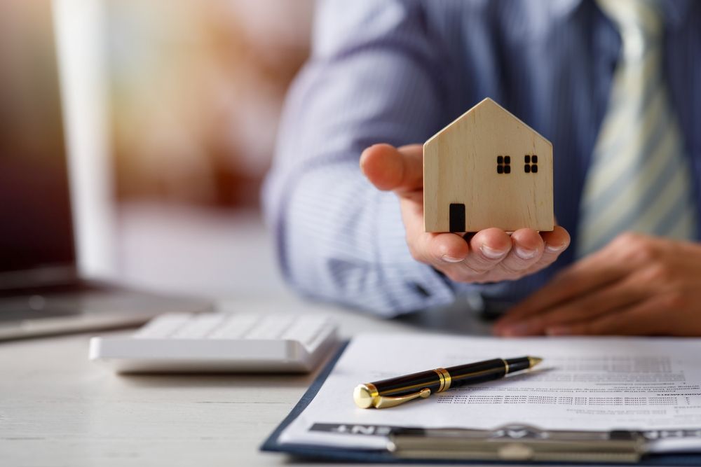 A person in a business shirt holds a small wooden house model over a desk with a contract and pen.