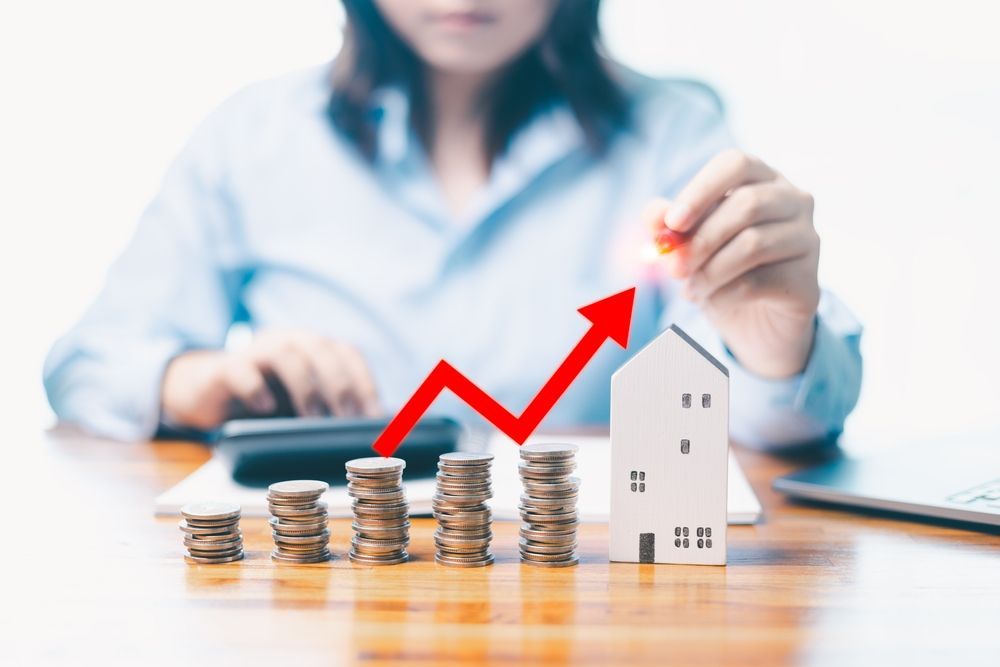 A person sits at a desk with stacks of coins and a small model house, with a digital red arrow indicating an upward trend.