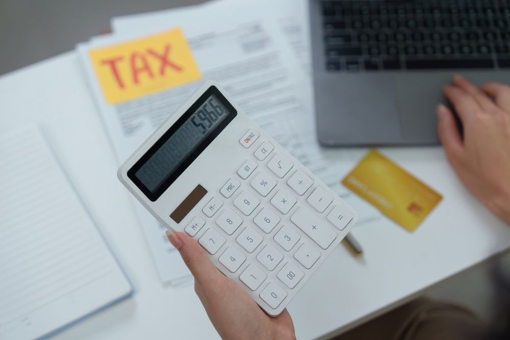 A person holds a calculator showing 59.66 near tax documents, a gold card, and a laptop on a white desk.