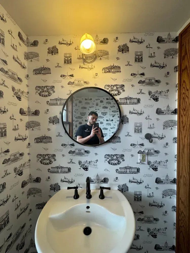 A person takes a mirror selfie in a bathroom with patterned black-and-white wallpaper and a round mirror.