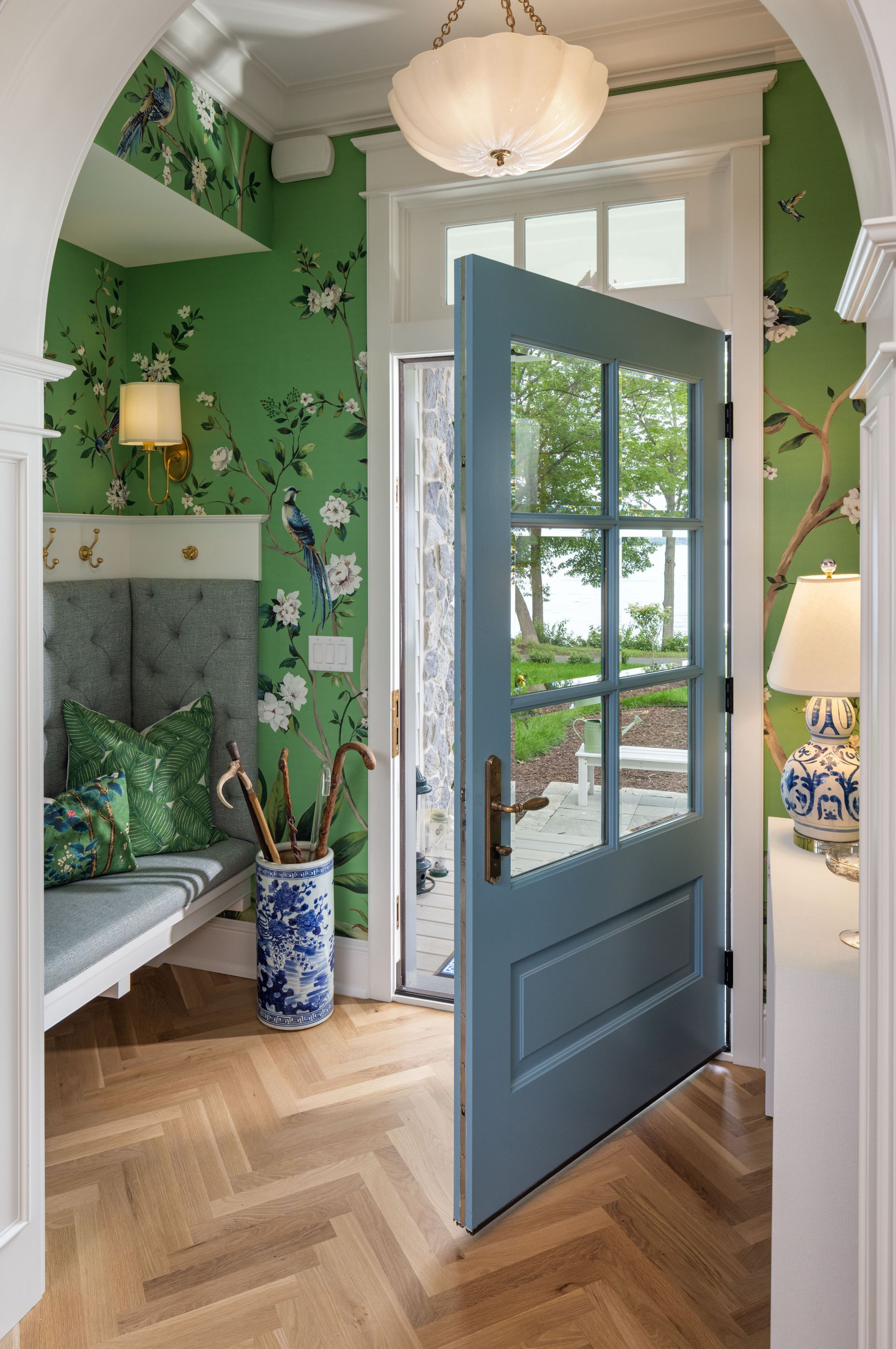 An open blue front door leads to an entryway with green floral wallpaper, a built-in bench, and herringbone wood floors.