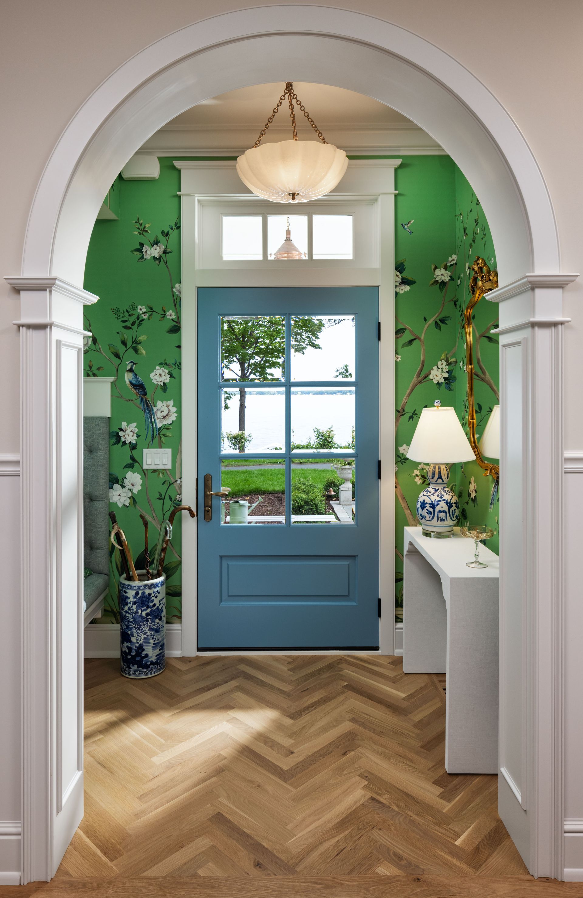 An arched doorway frames a foyer with green floral wallpaper, a blue door, herringbone wood floors, and a table lamp.