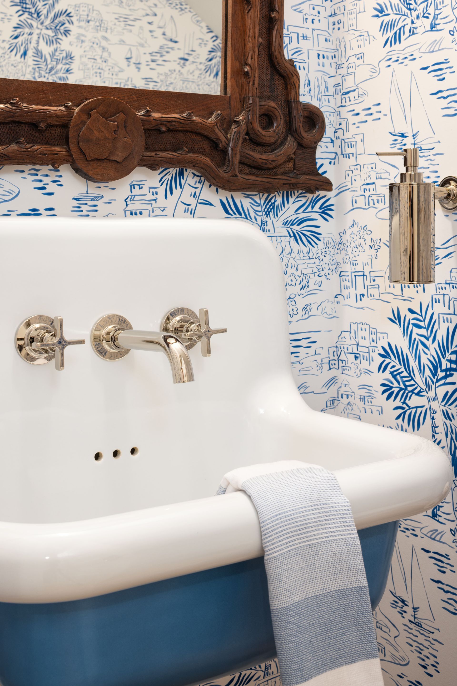A wall-mounted white and blue sink with silver fixtures against blue and white toile wallpaper, with a hand towel draped.