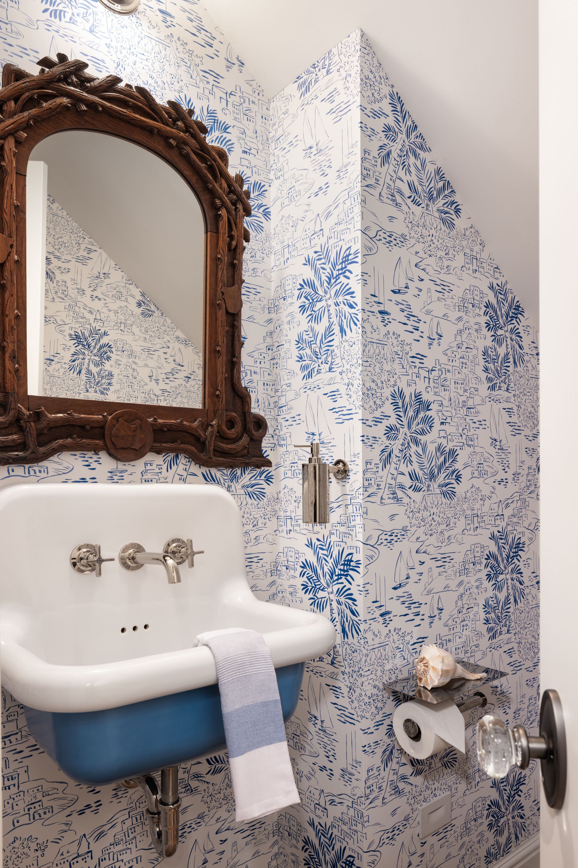 A wall-mounted blue and white sink sits below an ornate wooden mirror against blue floral wallpaper in a bathroom.
