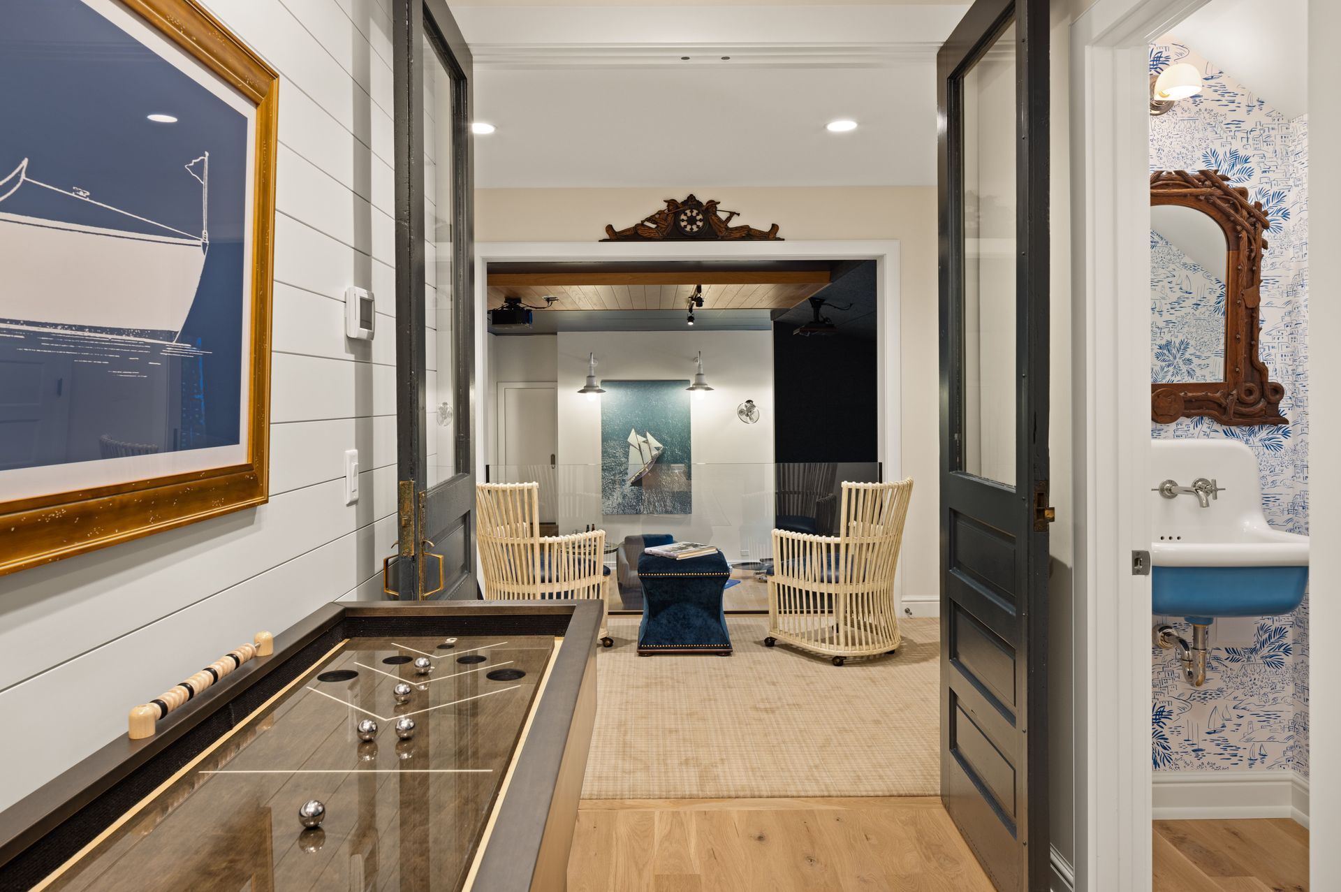 A shuffleboard table sits in a hallway, leading to a living area with two wicker chairs and a blue-and-white powder room.