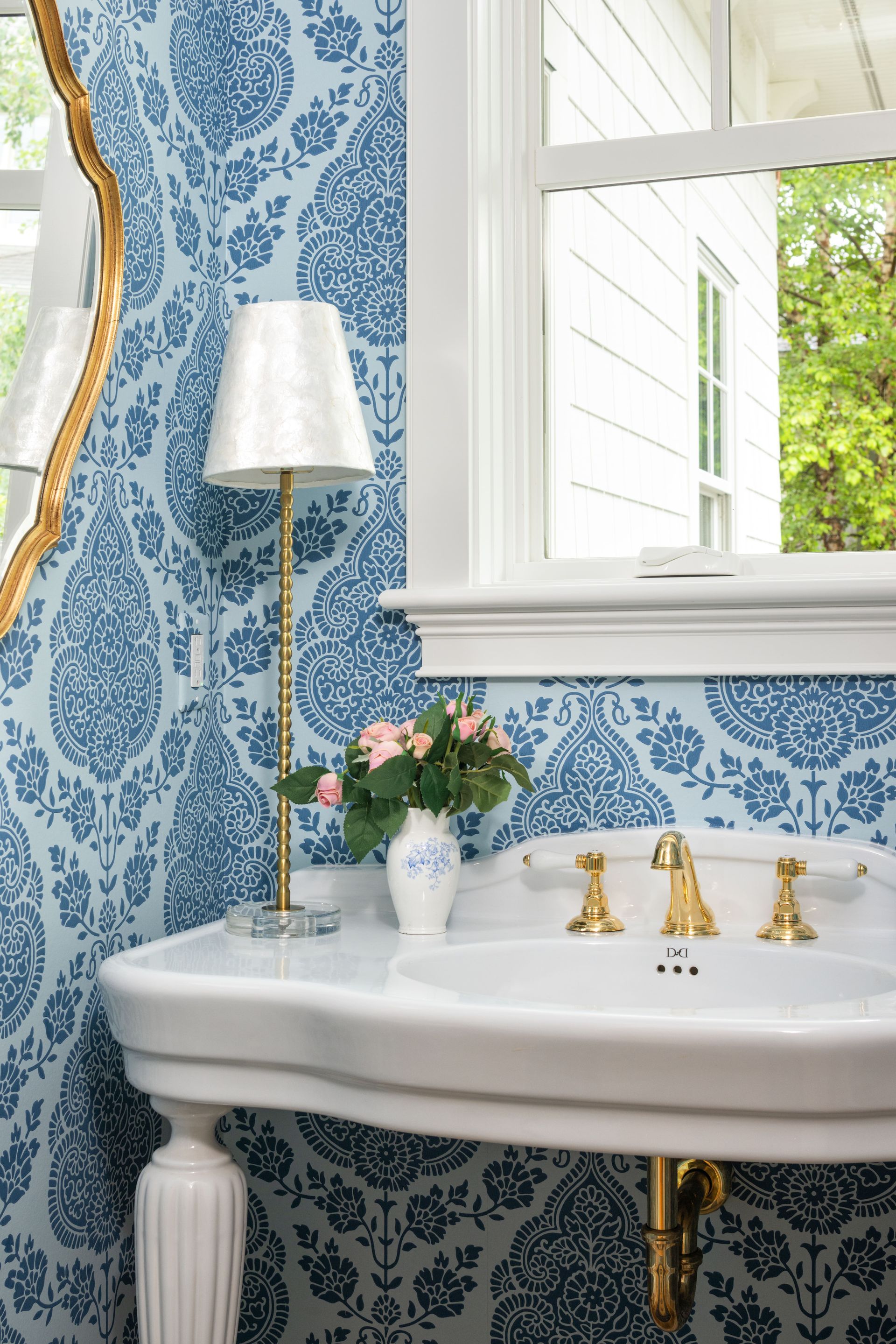 A bathroom sink with brass fixtures and a small floral lamp against blue and white damask patterned wallpaper.