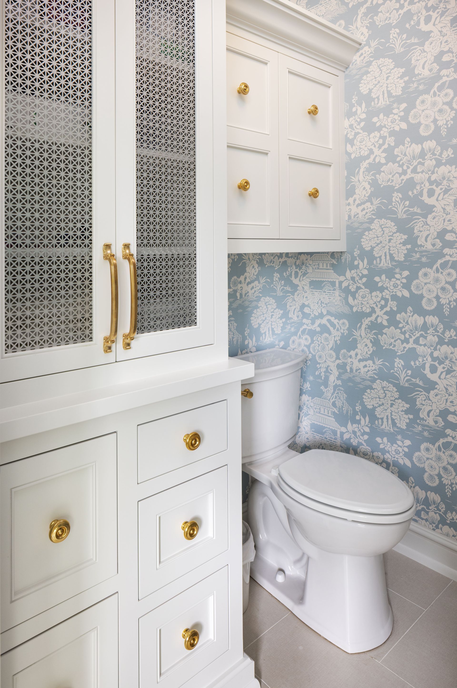 A bathroom with white cabinets featuring gold hardware, patterned glass, and blue and white floral wallpaper behind a toilet.