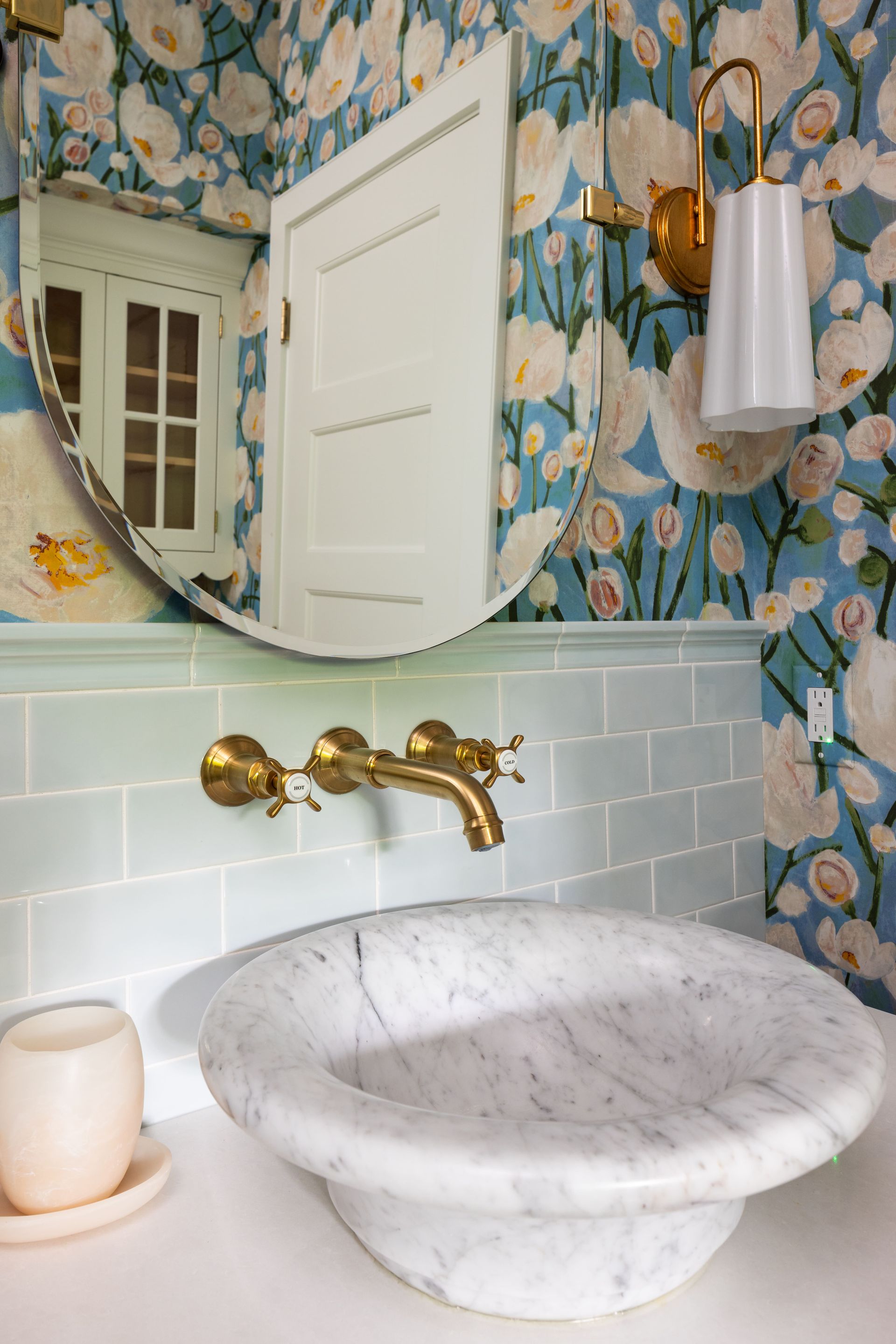 A marble vessel sink with gold fixtures, pale blue subway tile backsplash, and floral wallpaper reflecting in a mirror.