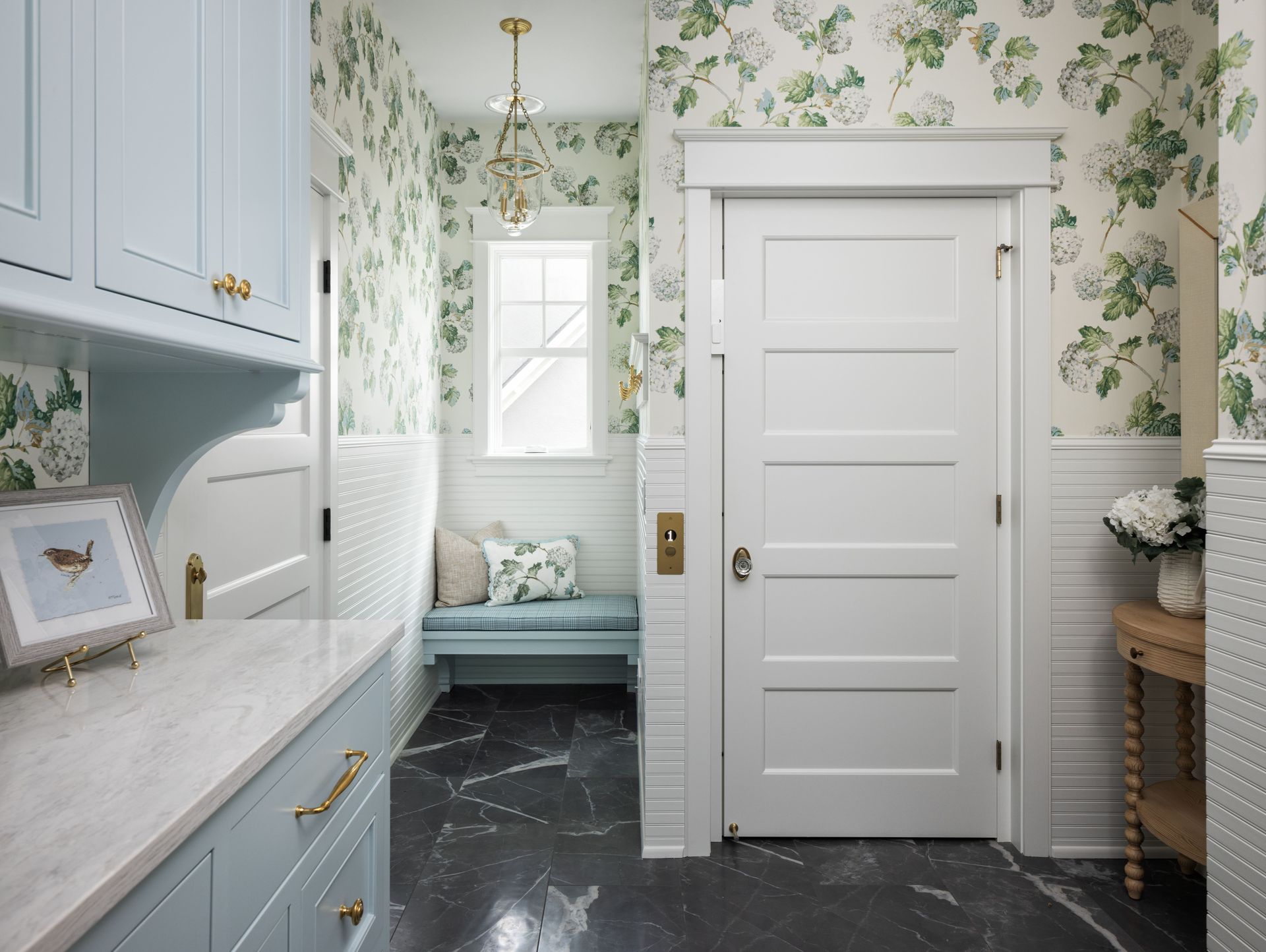 A bright entry hallway with light blue cabinets, floral wallpaper, white beadboard, a white door, and dark tile floors.
