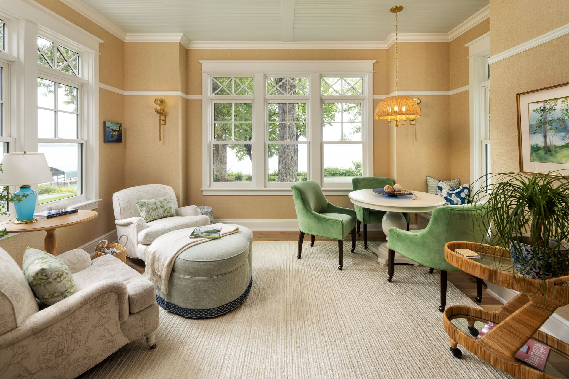 A sunlit living room with cream walls, beige armchairs, a large ottoman, and a round dining table with green chairs.