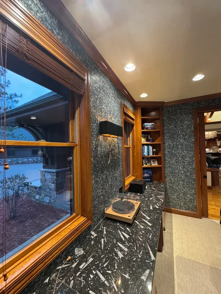A room with dark patterned wallpaper, a black marble countertop, a wooden-framed window, built-in shelves, and a lamp.