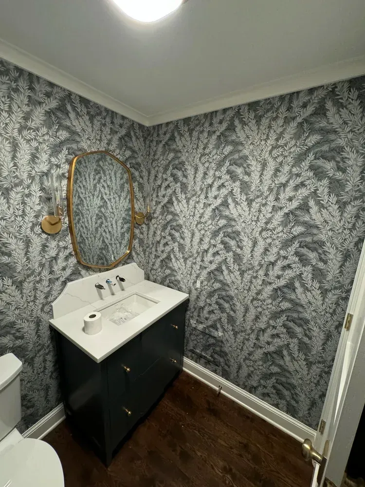 A small bathroom features a dark blue vanity with a white top, a gold-framed mirror, and leafy grey and white wallpaper.