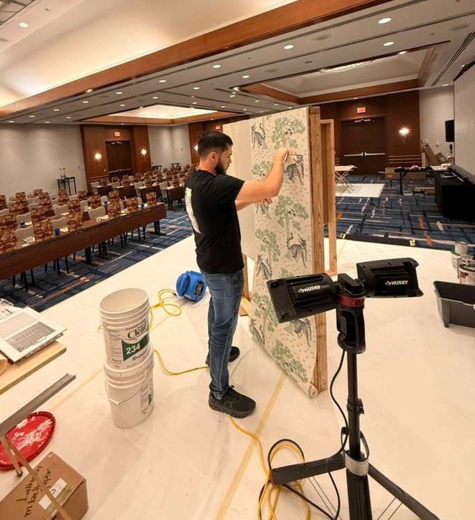 A person applies patterned wallpaper to a vertical wooden board inside a large, empty conference room.