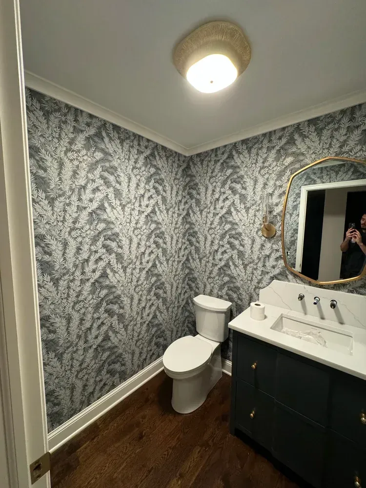 A bathroom with blue-and-white botanical wallpaper, a dark vanity, white countertop, and a modern gold-framed mirror.