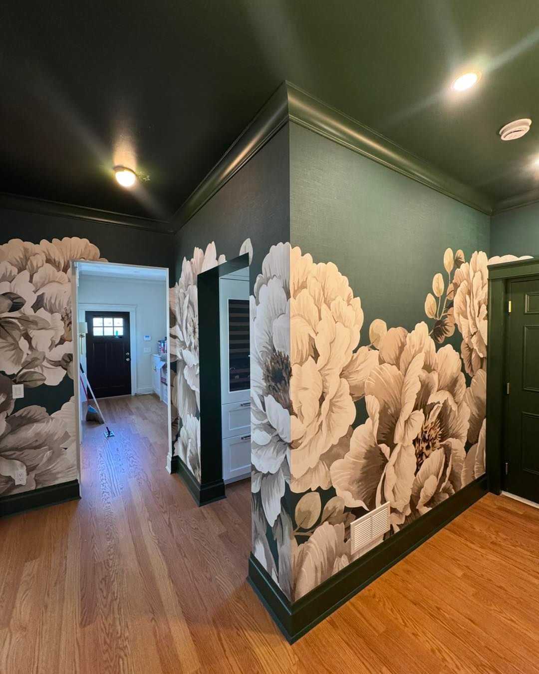 A hallway with dark green walls featuring a large-scale white floral mural, black trim, and light hardwood flooring.