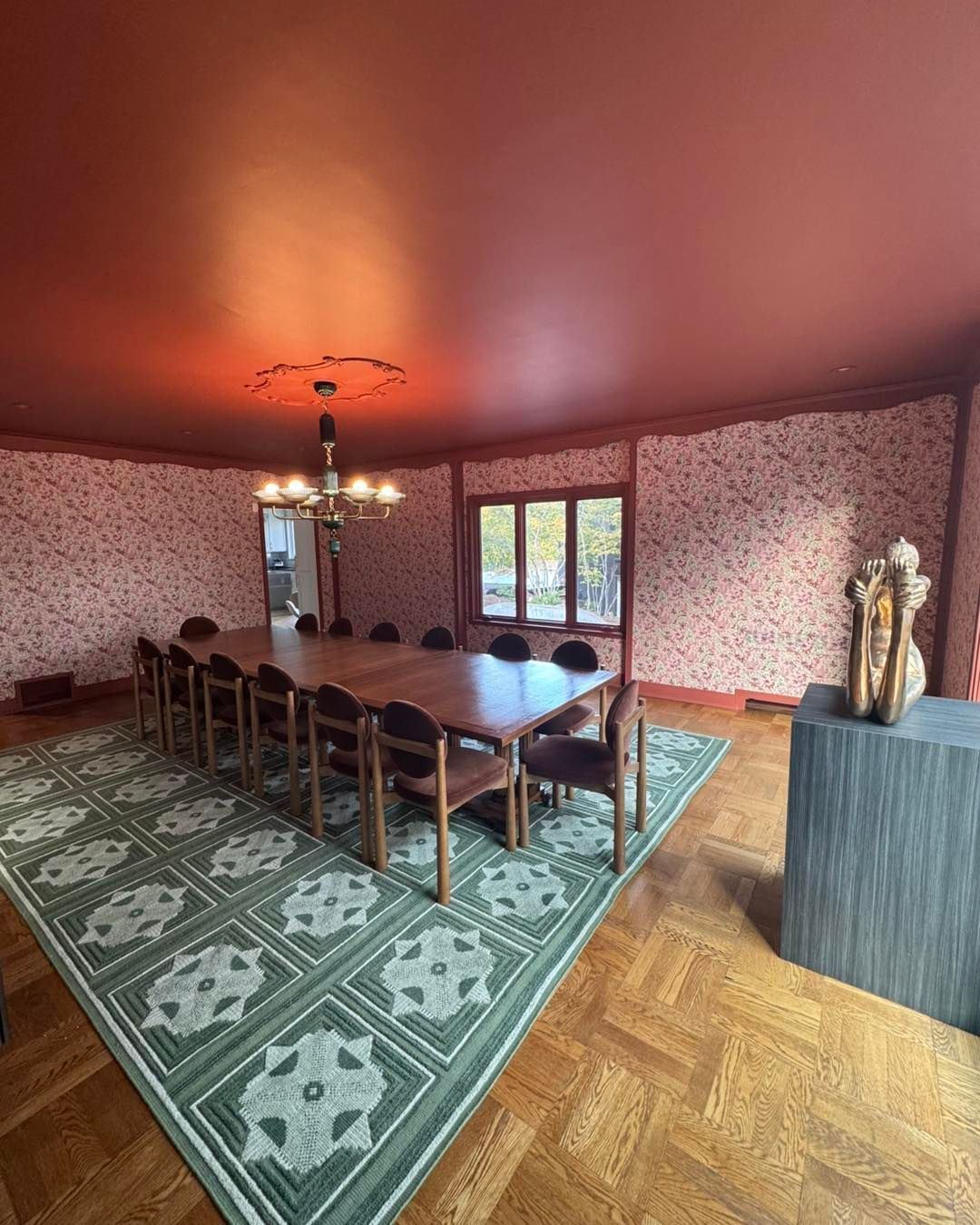 A formal dining room features a long wooden table, green patterned rug, floral wallpaper, and a terracotta ceiling.