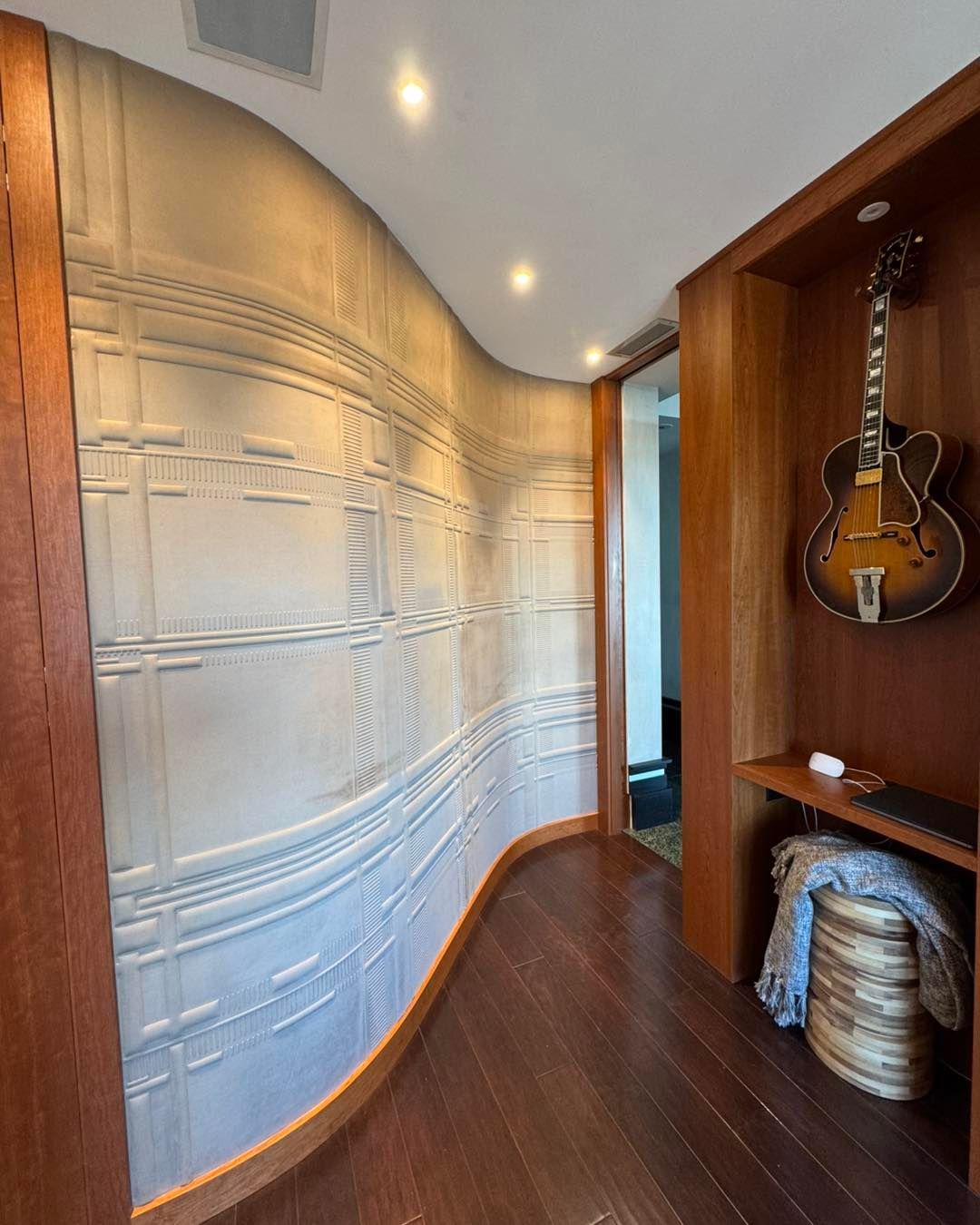 A curved wall with textured white panels, dark wooden floors, and a hollow alcove displaying a hanging guitar.