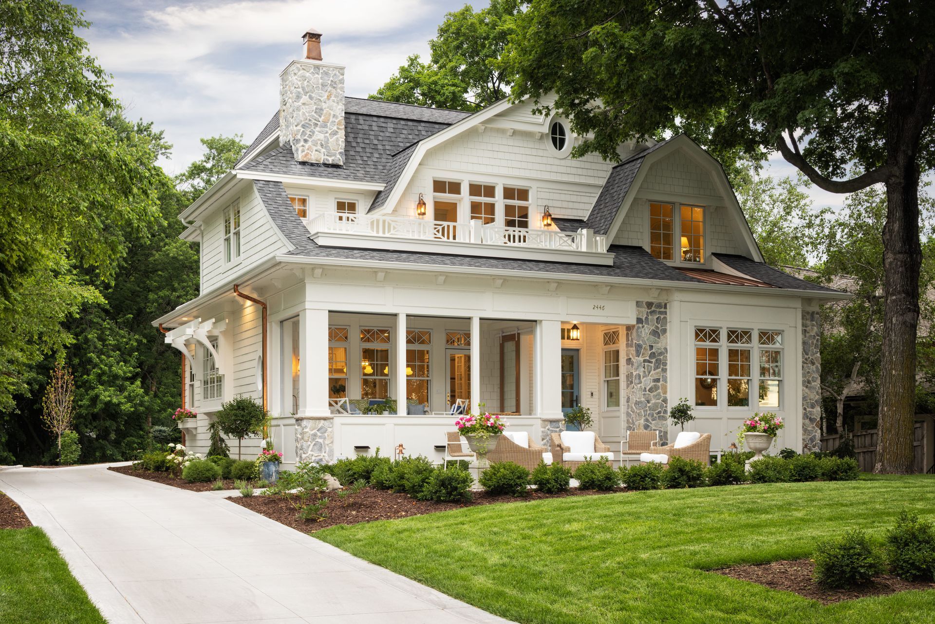 A white two-story craftsman-style house with a wraparound porch, stone accents, and a driveway set in a lush green yard.