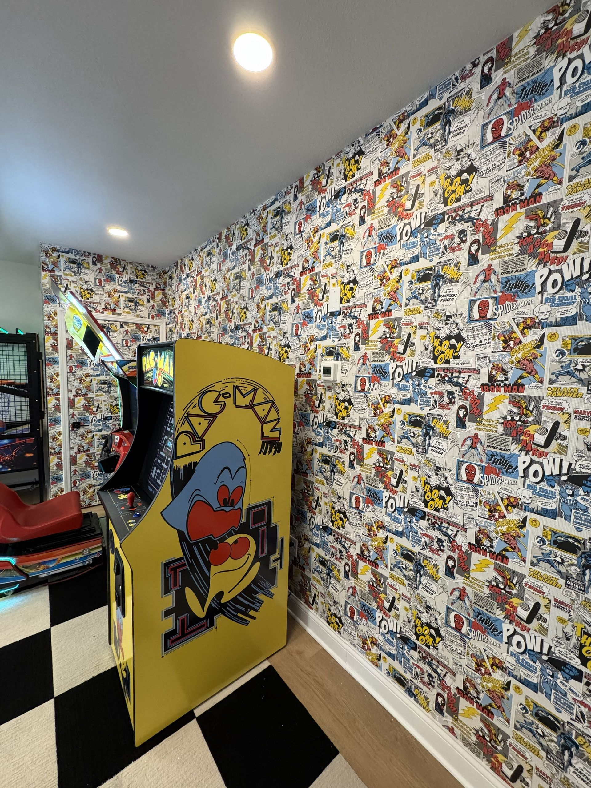 A yellow Pac-Man arcade cabinet stands in a room with black-and-white checkered floors and comic book wallpaper.