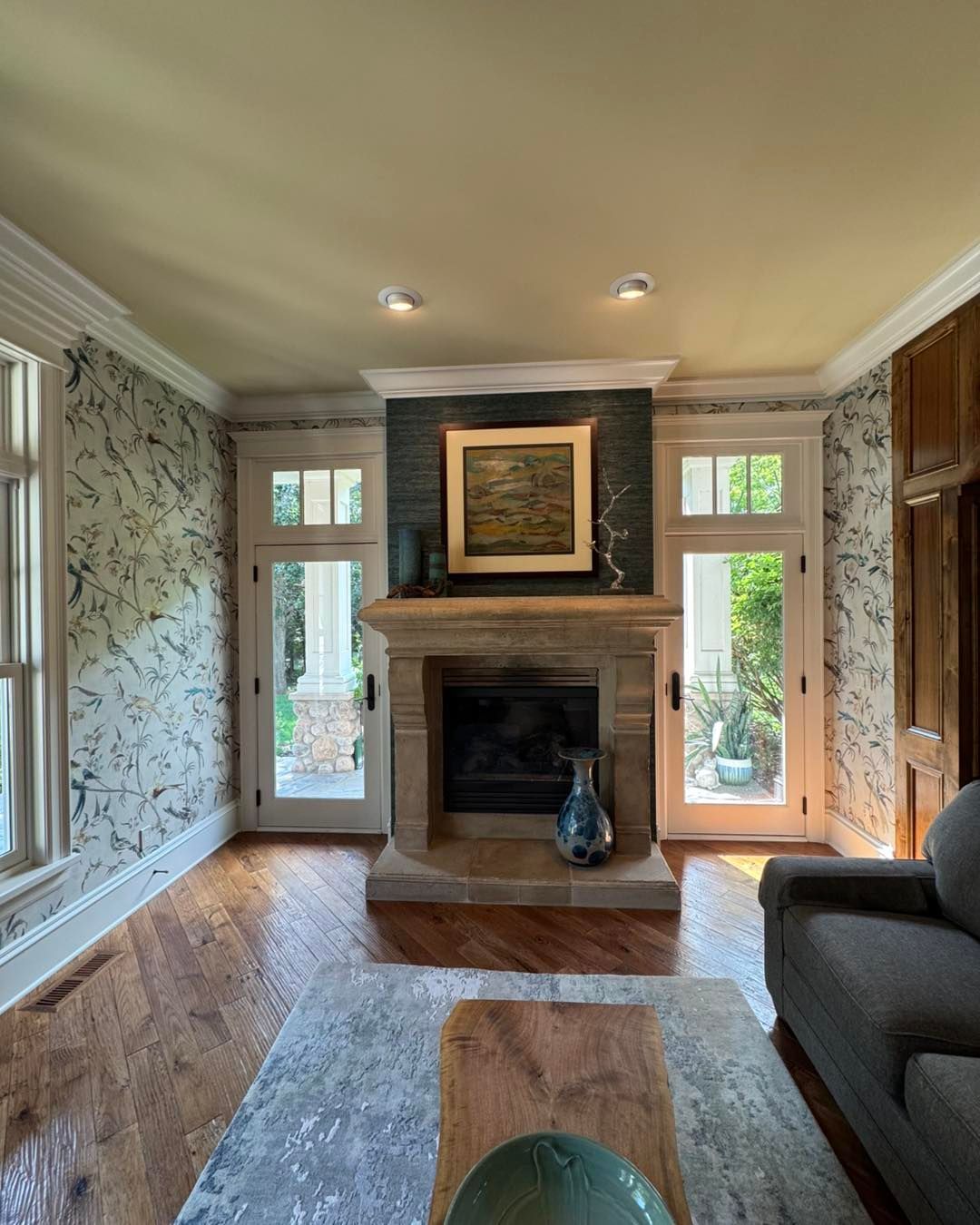 A fireplace with a stone mantel sits between two glass doors, accented by patterned wallpaper and a gray sofa.