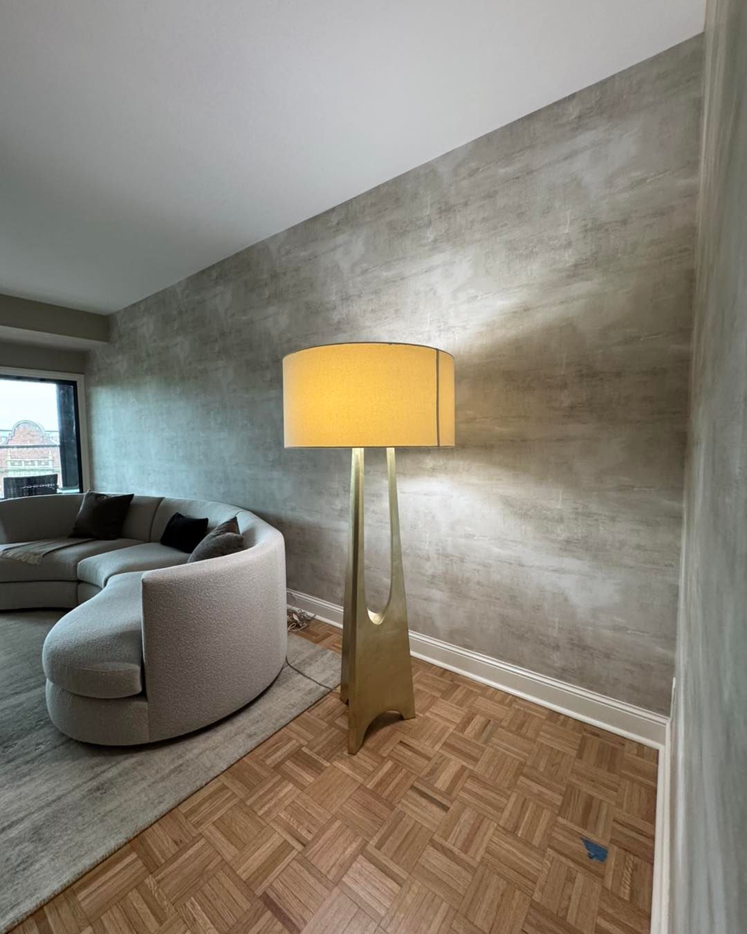 A tall gold floor lamp with a drum shade stands before a textured gray wall, next to a curved gray sofa on parquet floors.