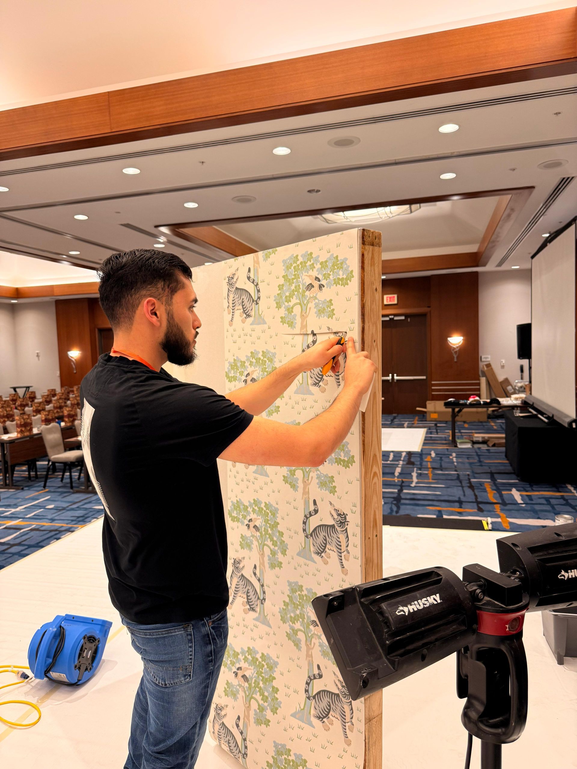 A person hangs patterned wallpaper on a wooden display board in a large, carpeted conference room.