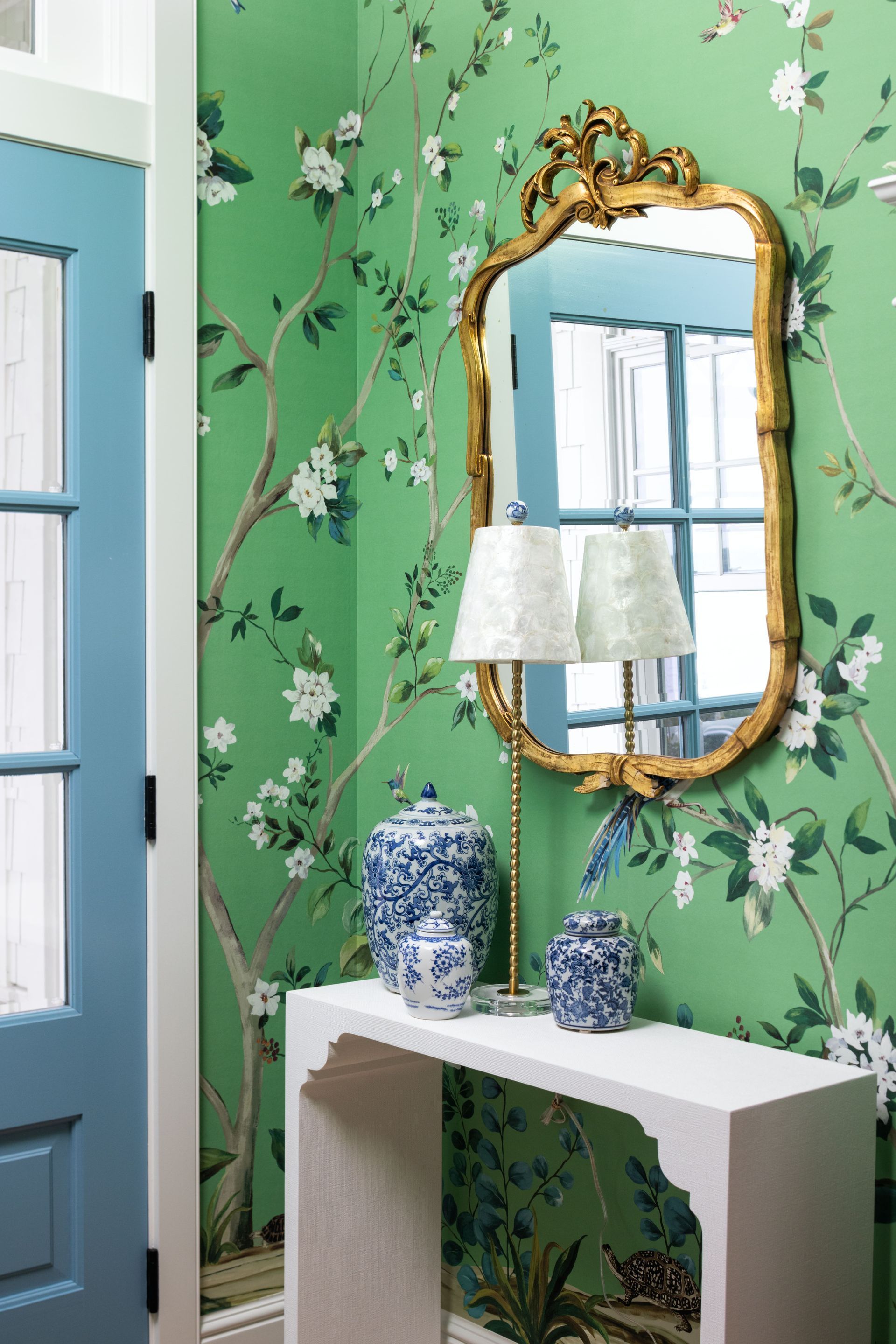 A white console table sits against a wall with green floral wallpaper, featuring blue-and-white vases and a gold mirror.