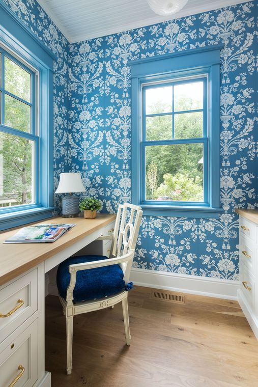 A small office nook with blue and white patterned wallpaper, a light wood desk, and a blue upholstered chair.