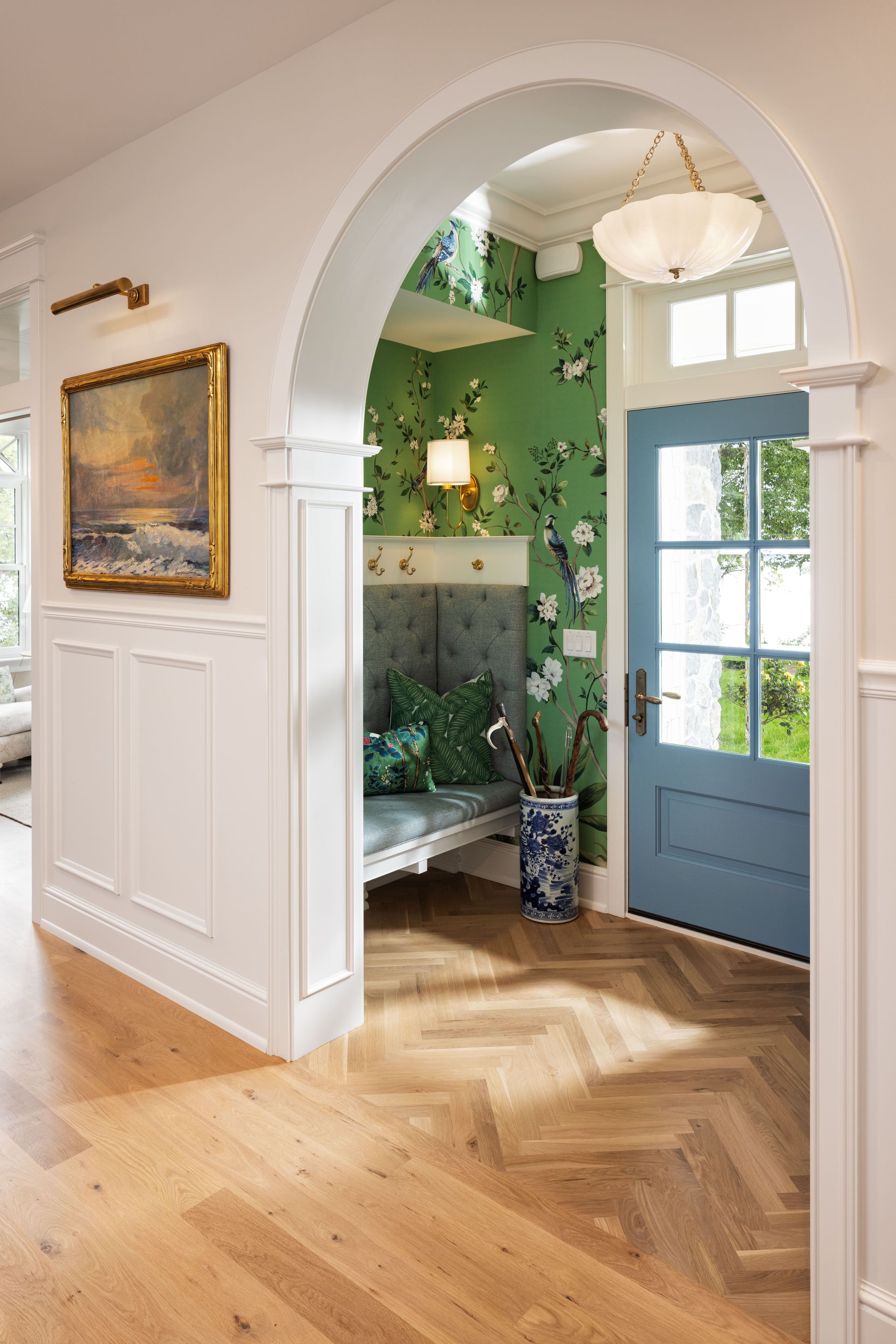 An arched doorway leads to a bright entryway with a blue front door, green floral wallpaper, and herringbone wood floors.