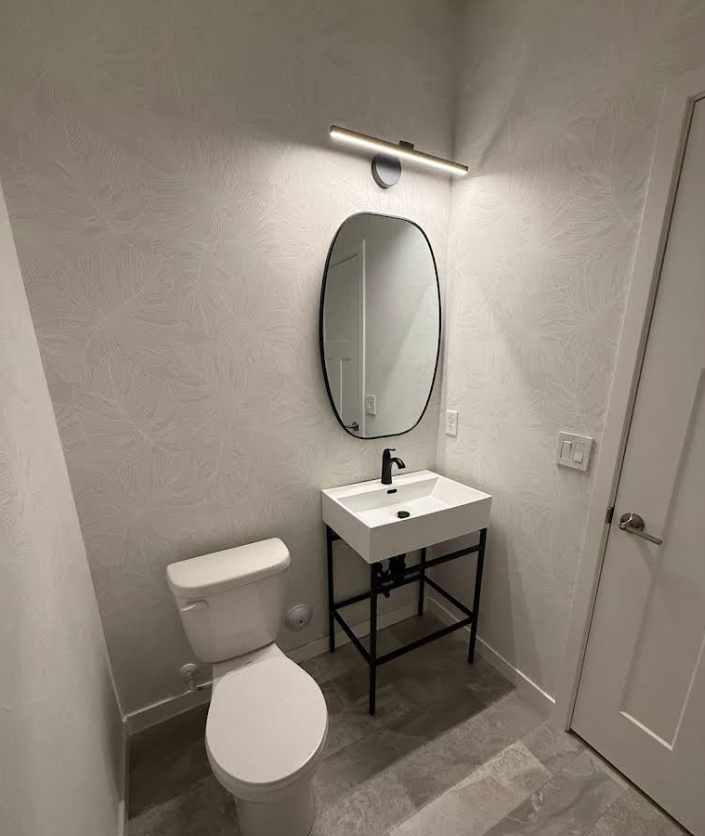 A minimalist half-bathroom with a white toilet, a modern rectangular sink on a black metal frame, and an oval mirror.