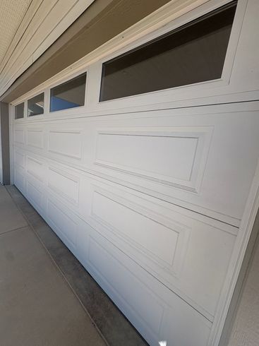 White paneled garage door with small windows along the top, viewed at an angle beside a concrete driveway