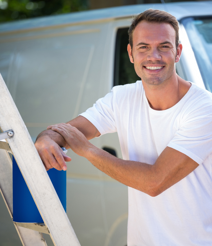 Man in white shirt smiles, leans on a ladder, van in the background.