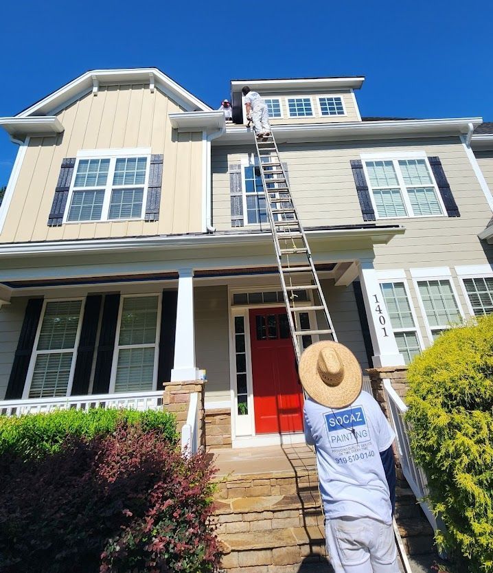 Two painters on a ladder, painting the trim of a two-story beige house with red door and blue sky.