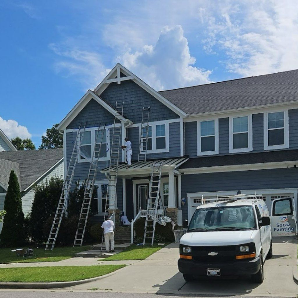 Painters on ladders work on the blue siding of a two-story house with a white work van parked in the driveway.