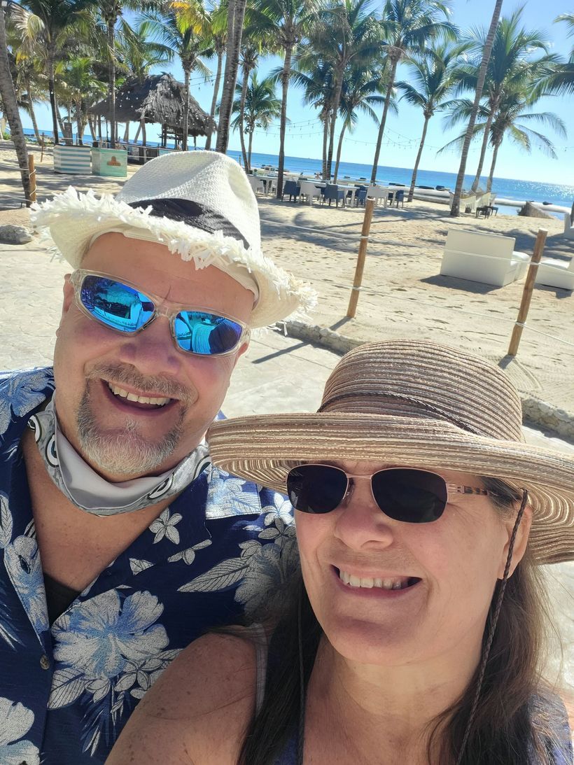 Woman in straw hat smiles, holds phone, stands before a tall ornate building with towers.
