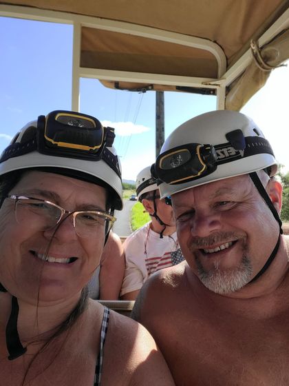 Two people smiling in a golf cart, wearing helmets with headlamps. Sunny outdoors.