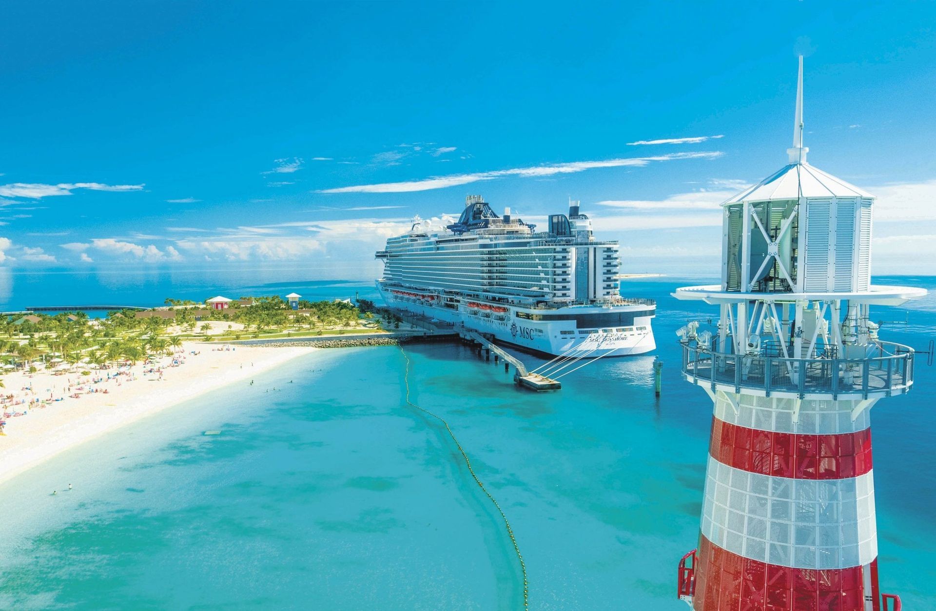 Cruise ship docked at a beach island with a red and white lighthouse, clear blue water and sky.